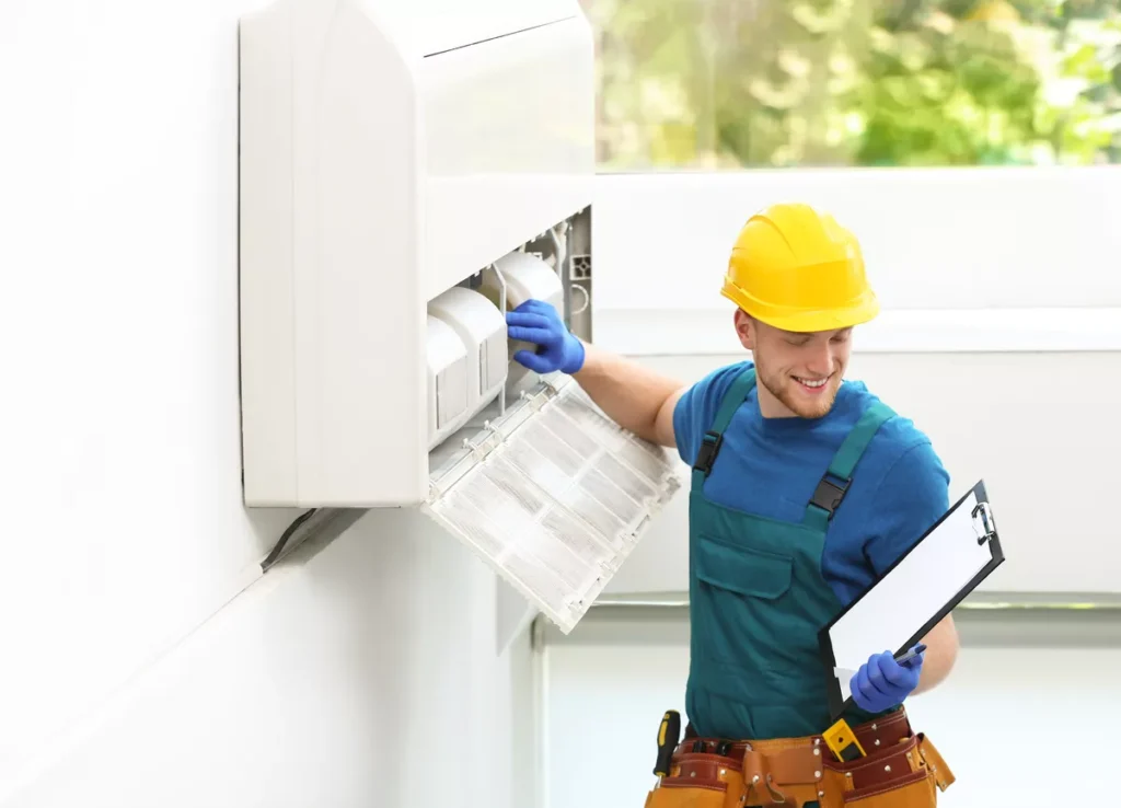 A smiling AC technician in a yellow hard hat and green overalls giving a successful service update while holding a clipboard in a Durham home.