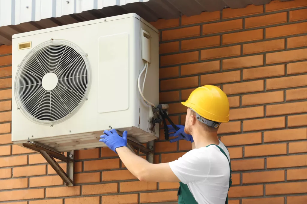 Safety-certified installation technician in a hardhat mounting a new air conditioning condenser unit to a brick exterior wall in Durham.