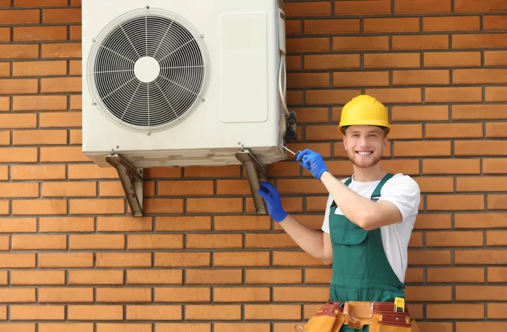 A smiling HVAC technician in a yellow hard hat and green overalls performing maintenance on an exterior AC unit against a brick wall in Durham.