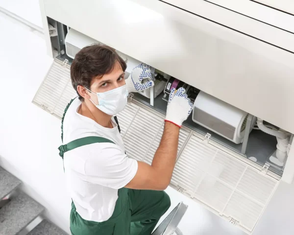 A professional Durham HVAC technician wearing a face mask and gloves while performing an indoor AC unit repair on a ladder.